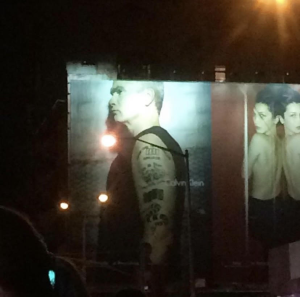 photograph of a billboard ad for Calvin Klein mounted on the side of a building in New York City, on the left is musician, author, comedian Henry Rollins wearing a black muscle tank, and on the right two models topless models shown from the side, wearing black skirts.